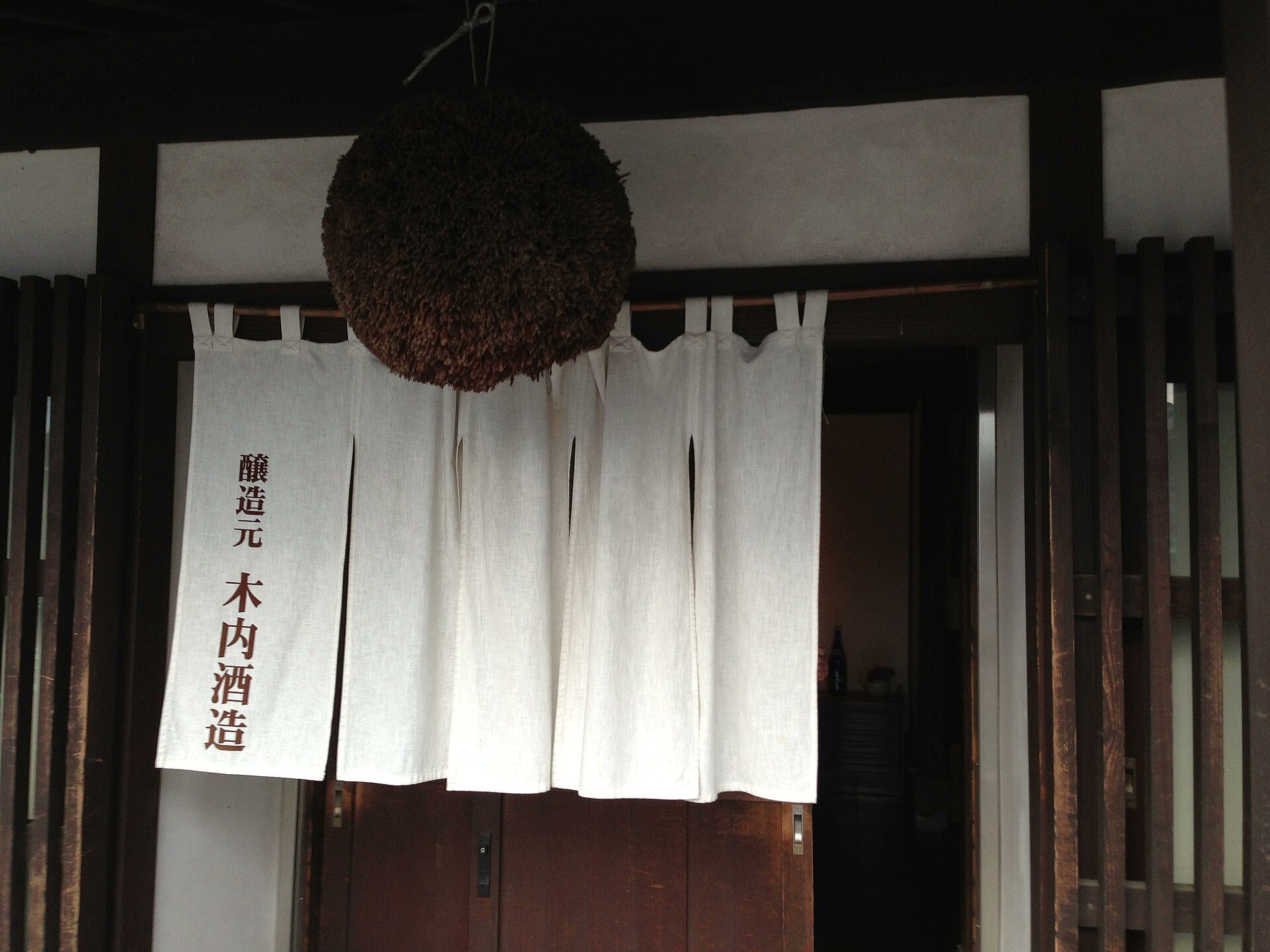 A noren curtain hung at a sake brewery entrance with brewery kanji, a cedar sugidama ball above